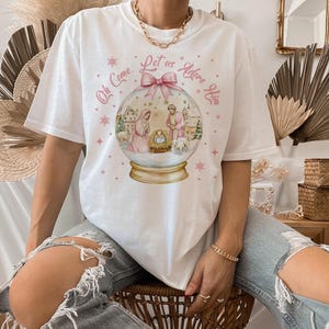K&ouml;nnte beinhalten: Wei&szlig;es T-Shirt mit einem rosa-wei&szlig;en Schneekugel-Design. Die Schneekugel zeigt eine Weihnachtskrippe mit Maria, Josef und dem Jesuskind. Der Text auf dem Shirt lautet "Oh Come, Let Us Adore Him".