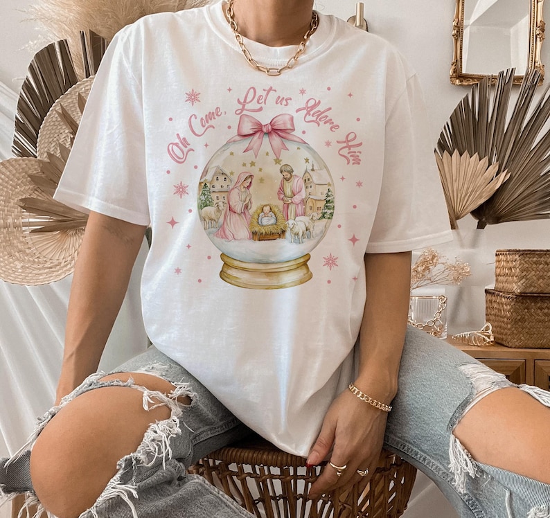 K&ouml;nnte beinhalten: Wei&szlig;es T-Shirt mit einem rosa-wei&szlig;en Schneekugel-Design. Die Schneekugel zeigt eine Weihnachtskrippe mit Maria, Josef und dem Jesuskind. Der Text auf dem Shirt lautet "Oh Come, Let Us Adore Him".
