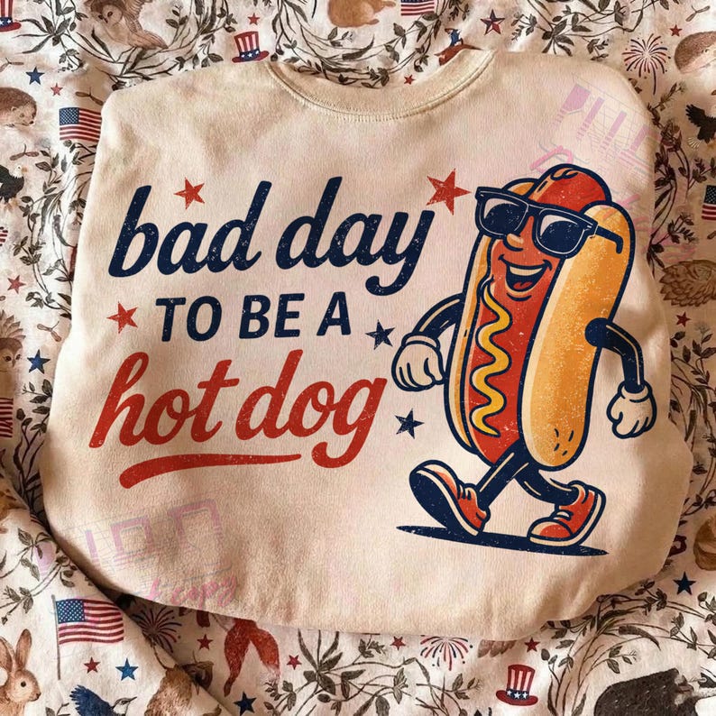 Puede incluir: Sudadera de color crema con el texto "bad day to be a hot dog" en azul marino y rojo. Un perrito caliente de dibujos animados con gafas de sol caminando. El dise&ntilde;o incluye estrellas.