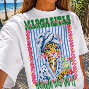 Pu&ograve; includere: T-shirt bianco con grafica colorata. La grafica presenta un leopardo con un accappatoio da spa e un cocktail, con le parole "Margaritas Made Me Do It" in testo verde e giallo. Lo sfondo ha strisce blu e bianche.