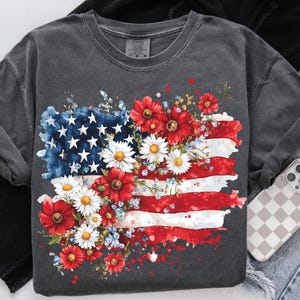 May include: A gray t-shirt with a watercolor design of an American flag with red, white, and blue flowers. The flag is faded and has a distressed look.