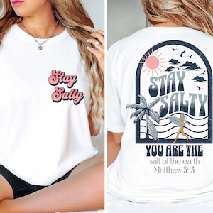 May include: White t-shirt with a retro-style graphic on the front that says "Stay Salty". The back of the shirt features a beach scene with a palm tree, sun, and waves. The text "Stay Salty" is also on the back, along with the words "You are the salt of the earth Matthew 5:13".
