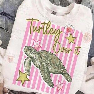 May include: White sweatshirt with a graphic of a sea turtle, pink and white striped background, and the text "Turley Over it". The design includes yellow stars and pink stars.
