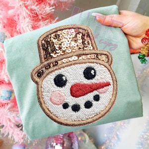 Puede incluir: Una camiseta verde claro con un aplique de cara de muñeco de nieve. El muñeco de nieve tiene un sombrero con lentejuelas, ojos negros, una nariz de zanahoria roja y una mejilla rosa. El aplique está delineado con hilo dorado.