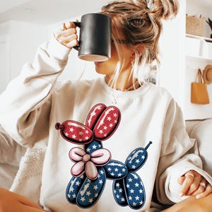 May include: A white sweatshirt with a red, white, and blue balloon animal design. The balloon animal is shaped like a dog and has stars on it.
