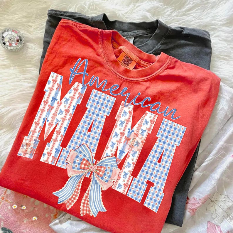 Op de afbeelding: Een rood t-shirt met de woorden "American MAMA" in blauwe letters en blokletters. De blokletters zijn versierd met een ruitpatroon en strikken in rood, wit en blauw. Een grijs t-shirt is eronder zichtbaar.
