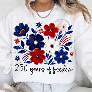 May include: White sweatshirt featuring a floral design in red, white, and blue. The design includes stylized flowers, leaves, and the text "250 years of freedom" in a script font. The sweatshirt has a crew neck.