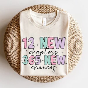 K&ouml;nnte beinhalten: Ein wei&szlig;es T-Shirt mit dem Text "12 New Chapters 365 New Chances" in rosa, gr&uuml;n und lila Schrift.