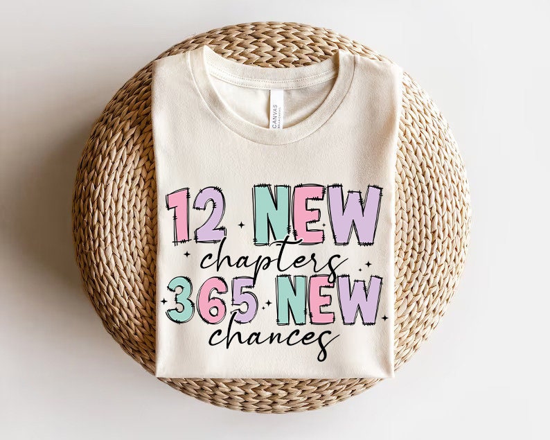 K&ouml;nnte beinhalten: Ein wei&szlig;es T-Shirt mit dem Text "12 New Chapters 365 New Chances" in rosa, gr&uuml;n und lila Schrift.