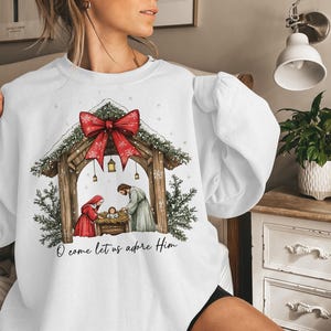 May include: White sweatshirt featuring a Nativity scene with a red bow and the text "O come let us adore Him." The design includes a wooden stable, figures in red and gray, and evergreen accents. A festive holiday-themed garment.
