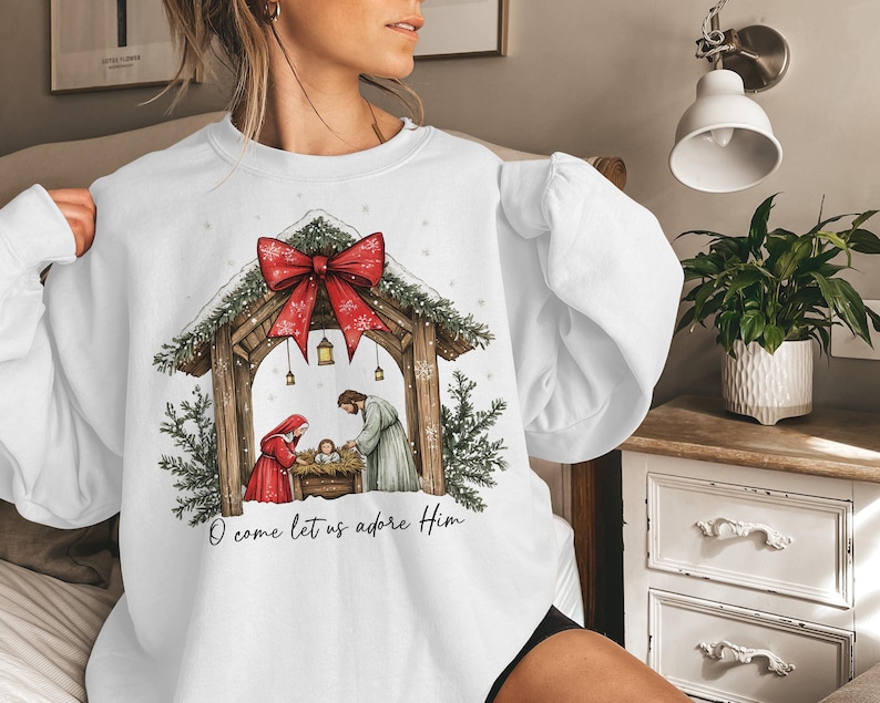 May include: White sweatshirt featuring a Nativity scene with a red bow and the text "O come let us adore Him." The design includes a wooden stable, figures in red and gray, and evergreen accents. A festive holiday-themed garment.