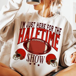 May include: White sweatshirt with red text that reads "I'm just here for the halftime show" with a football and two red helmets.