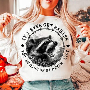 May include: White sweatshirt featuring a black and white illustration of a raccoon with an open mouth, surrounded by barbed wire and text that reads "IF I EVER GET RABIES YOU'RE HIGH ON MY BITIN' LIST."