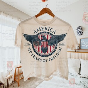 May include: Cream-colored t-shirt with a distressed graphic featuring an eagle with outstretched wings, a shield, and the text "AMERICA 1776-2026 250 YEARS OF FREEDOM". The design is in shades of blue, red, and white.
