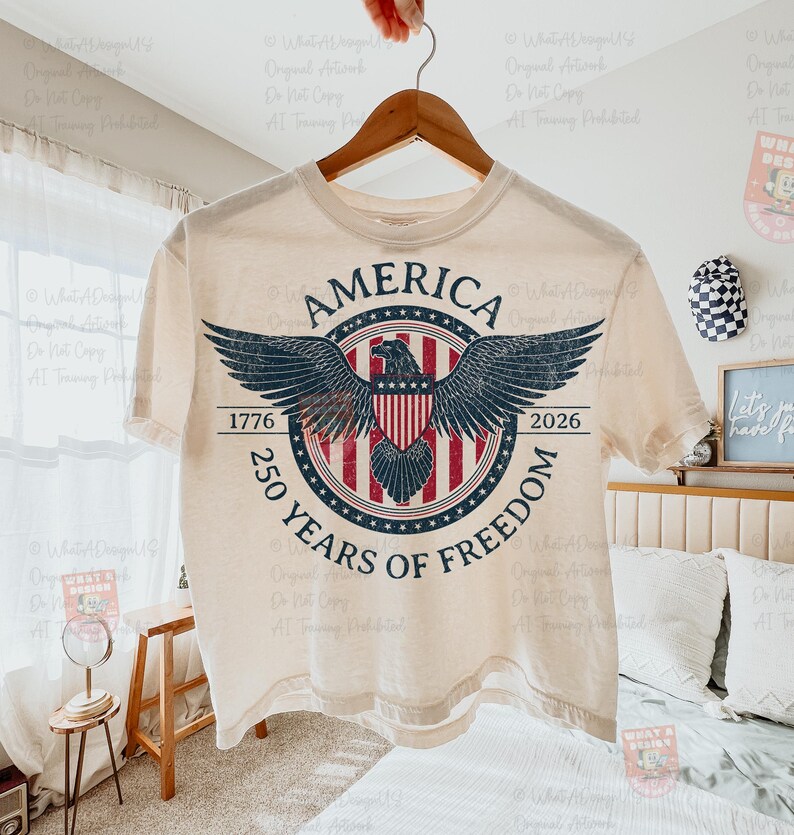 May include: Cream-colored t-shirt with a distressed graphic featuring an eagle with outstretched wings, a shield, and the text "AMERICA 1776-2026 250 YEARS OF FREEDOM". The design is in shades of blue, red, and white.