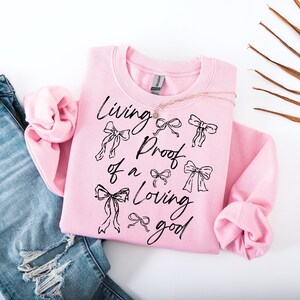 May include: A pink sweatshirt with the text "Living proof of a loving good" in black lettering. The text is surrounded by small black bow designs.