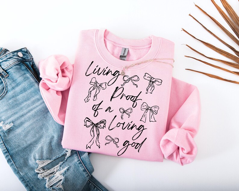 May include: A pink sweatshirt with the text "Living proof of a loving good" in black lettering. The text is surrounded by small black bow designs.