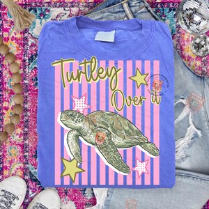 May include: A periwinkle blue t-shirt with the phrase "Turtley Over It" in a whimsical font. The design features a green sea turtle, pink and blue stripes, and yellow stars. A fun, casual top.