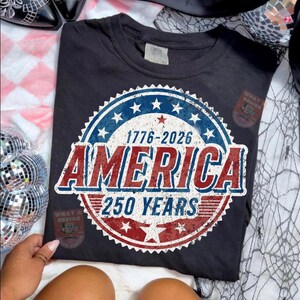 May include: Dark gray t-shirt with a patriotic design. The design features a circular emblem with the text "1776-2026 AMERICA 250 YEARS" in red, white, and blue. The shirt is a comfortable, casual style.