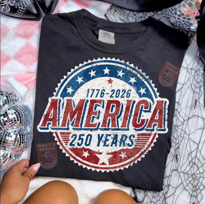 May include: Dark gray t-shirt with a patriotic design. The design features a circular emblem with the text "1776-2026 AMERICA 250 YEARS" in red, white, and blue. The shirt is a comfortable, casual style.