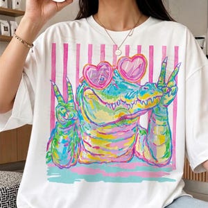 May include: White t-shirt featuring a colorful alligator wearing heart-shaped sunglasses and making peace signs. The alligator is painted in vibrant hues of green, yellow, pink, and blue. Pink vertical stripes and text are in the background.