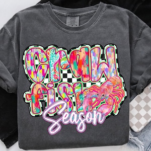 May include: A gray t-shirt with a colorful graphic design that says "Crawfish Season". The graphic features a pink crawfish and a checkered background.