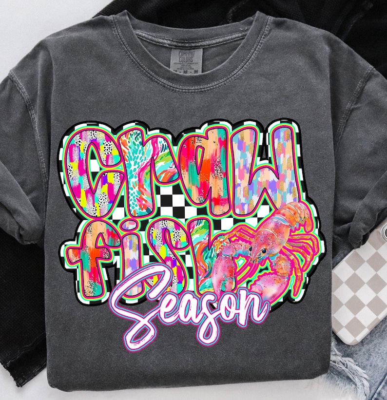 May include: A gray t-shirt with a colorful graphic design that says "Crawfish Season". The graphic features a pink crawfish and a checkered background.