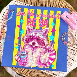 Puede incluir: Camiseta azul con un gráfico colorido de un mapache con gafas de sol y una bebida. El texto "See Ya wouldn't BE YA" está en rosa y amarillo. El mapache está en tonos de rosa, morado y blanco.