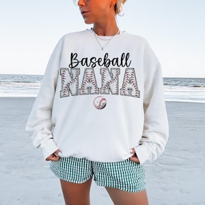 May include: White sweatshirt with the text "Baseball Nana" in black lettering. The letters are outlined with a baseball stitch design. A small baseball graphic is below the text.