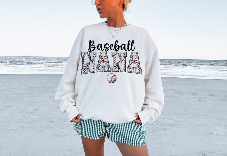 May include: White sweatshirt with the text "Baseball Nana" in black lettering. The letters are outlined with a baseball stitch design. A small baseball graphic is below the text.