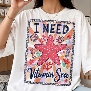 May include: White t-shirt with a colorful graphic featuring a red starfish, seashells, seahorses, and the text "I NEED Vitamin Sea." The design is framed with a rope-like border and a retro sunburst background.