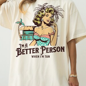 May include: A white t-shirt with a vintage-style graphic of a woman in a swimsuit and palm trees. The text "I'm a Better Person When I'm Tan" is printed below the graphic.