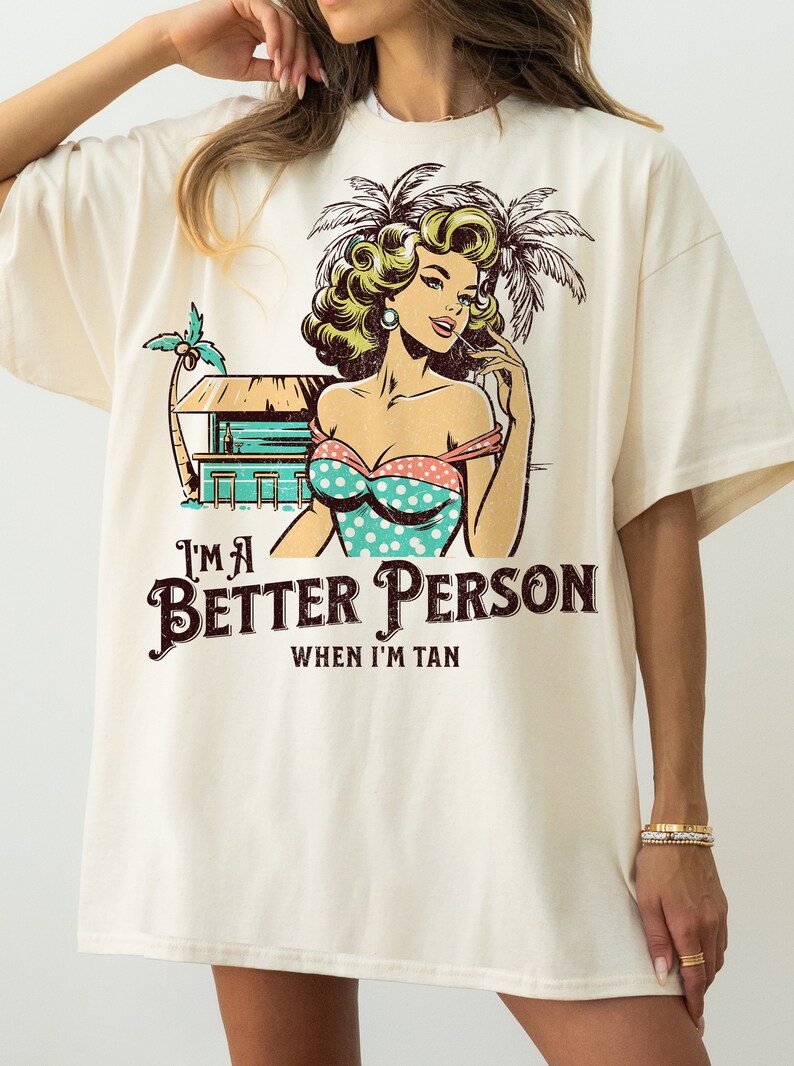 May include: A white t-shirt with a vintage-style graphic of a woman in a swimsuit and palm trees. The text "I'm a Better Person When I'm Tan" is printed below the graphic.