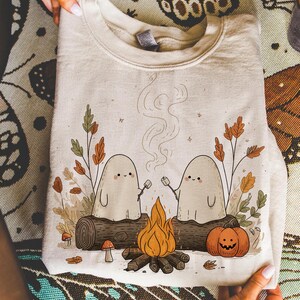 May include: Cream-colored sweatshirt featuring a campfire scene with two cartoon ghosts roasting marshmallows. The design includes a bonfire, a carved pumpkin, and autumn foliage. A cozy, fall-themed graphic.