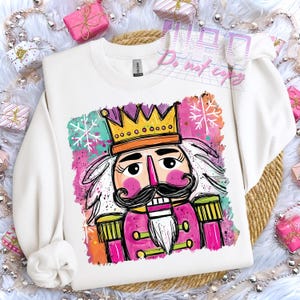 May include: White crewneck sweatshirt featuring a colorful illustration of a nutcracker soldier. The design includes a gold crown, pink uniform, and a black mustache. The background has snowflakes and splashes of color.