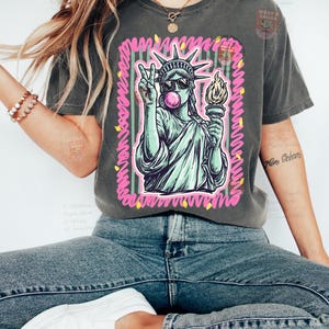 Puede incluir: Camiseta gris oscuro con un gr&aacute;fico de la Estatua de la Libertad con gafas de sol y haciendo una burbuja de chicle, con un signo de la paz. El dise&ntilde;o est&aacute; enmarcado con detalles en rosa y verde. La camiseta se combina con vaqueros azules.