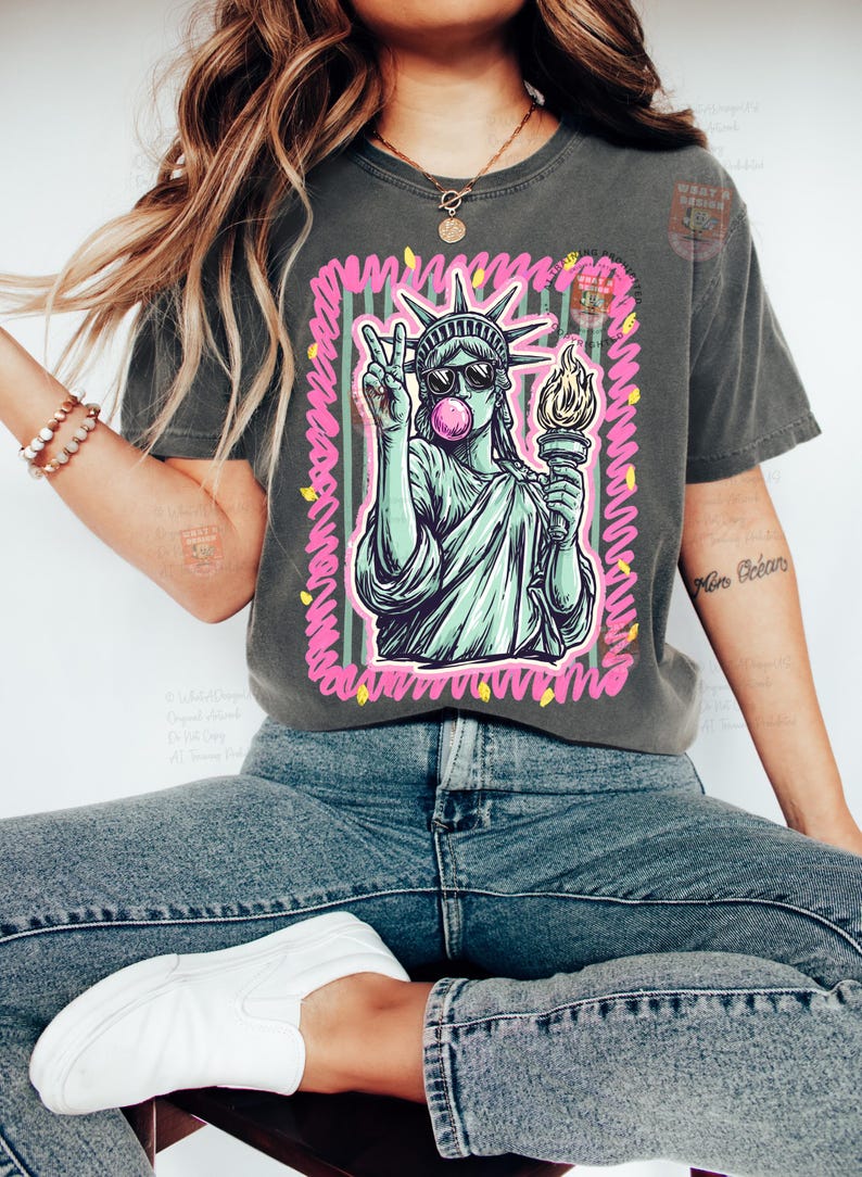 Puede incluir: Camiseta gris oscuro con un gr&aacute;fico de la Estatua de la Libertad con gafas de sol y haciendo una burbuja de chicle, con un signo de la paz. El dise&ntilde;o est&aacute; enmarcado con detalles en rosa y verde. La camiseta se combina con vaqueros azules.