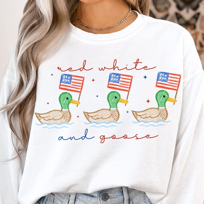 May include: White sweatshirt with a whimsical design featuring three ducks holding American flags. The text "red white and goose" is written in red and blue script, creating a patriotic theme.