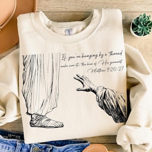 May include: A beige sweatshirt with a black and white illustration of a person reaching out a hand to another person. The text on the sweatshirt reads "If you're hanging by a thread make sure it's the hem of His garment. Matthew 9:20-27".