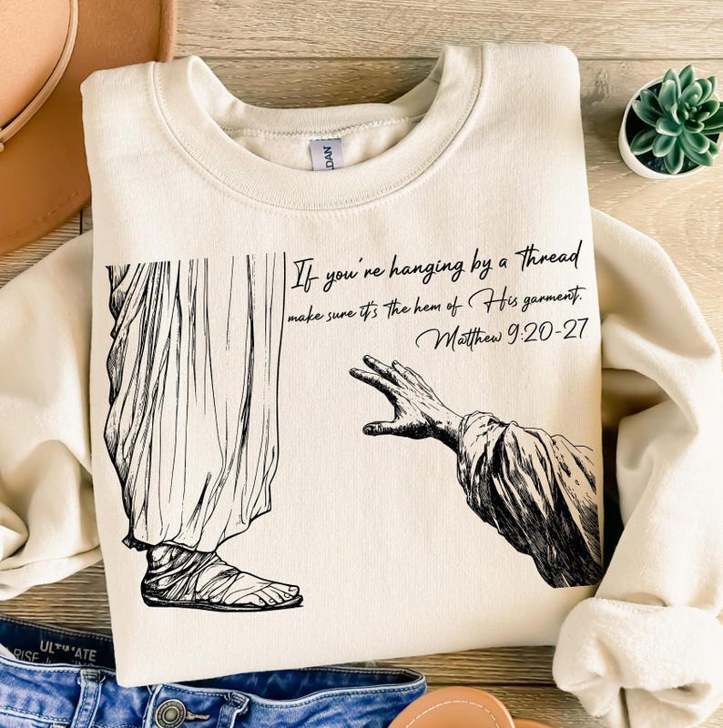 May include: A beige sweatshirt featuring a black and white illustration of a person reaching out a hand to another person. The text on the sweatshirt reads "If you're hanging by a thread make sure it's the hem of His garment. Matthew 9:20-27".