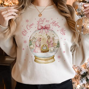 K&ouml;nnte beinhalten: Ein wei&szlig;er Pullover mit einem rosa-wei&szlig;en Schneekugel-Design. Die Schneekugel zeigt eine Weihnachtskrippe mit Maria, Josef und dem Jesuskind. Der Text auf dem Pullover lautet "Oh Come, Let Us Adore Him".