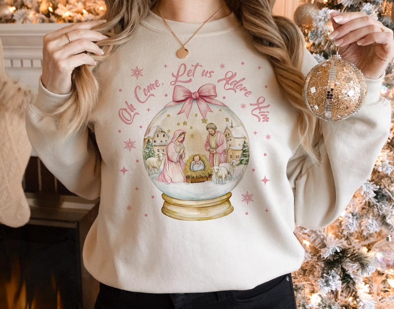 K&ouml;nnte beinhalten: Ein wei&szlig;er Pullover mit einem rosa-wei&szlig;en Schneekugel-Design. Die Schneekugel zeigt eine Weihnachtskrippe mit Maria, Josef und dem Jesuskind. Der Text auf dem Pullover lautet "Oh Come, Let Us Adore Him".