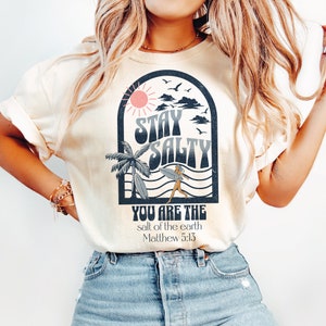 May include: A cream colored t-shirt with a graphic of a palm tree, a surfer, and the text "Stay Salty" in a retro font. The graphic is surrounded by a blue arch and waves. Below the graphic is the text "You are the salt of the earth Matthew 5:13".