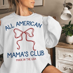 May include: White sweatshirt with the text "ALL AMERICAN MAMA'S CLUB" in blue and red, featuring a red bow graphic. The words "MADE IN THE USA" are printed below the club name.