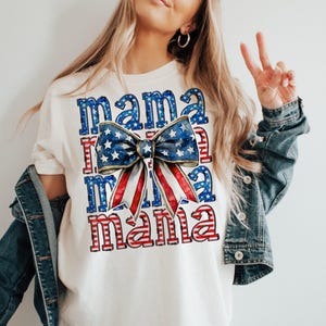 May include: Cream-colored t-shirt with the word "Mama" in red and blue, with a bow design featuring a blue star pattern and red and white stripes. The shirt is part of a patriotic theme.