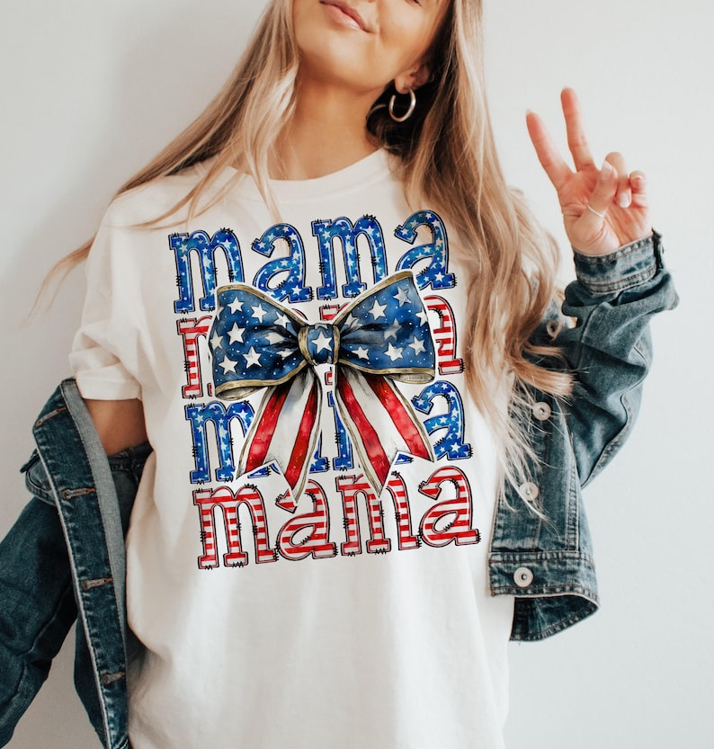 May include: Cream-colored t-shirt with the word "Mama" in red and blue, with a bow design featuring a blue star pattern and red and white stripes. The shirt is part of a patriotic theme.