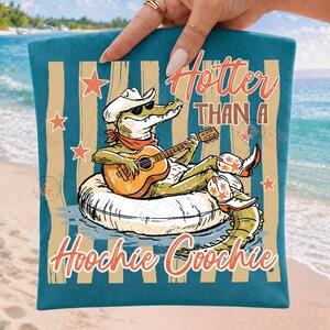 May include: Teal beach towel with a cartoon alligator wearing a cowboy hat and sunglasses, playing a guitar while floating on an inner tube. The text "Hotter Than A Hoochie Coochie" is displayed in orange.