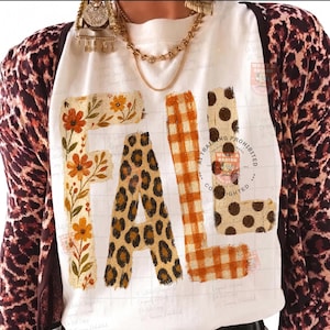 May include: A white t-shirt featuring the word FALL in large block letters. The F is decorated with orange flowers, the A has a leopard print, the L features an orange gingham pattern, and the second L has brown polka dots. A leopard print cardigan is worn over the shirt.
