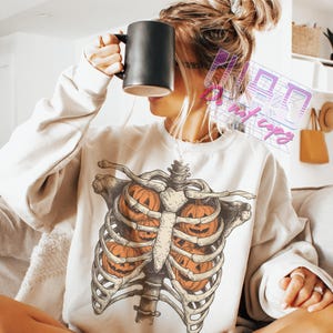 May include: Cream-colored sweatshirt featuring a skeletal rib cage design with three carved orange pumpkins inside. The graphic is detailed with shading and highlights. The sweatshirt is a casual, long-sleeved garment.