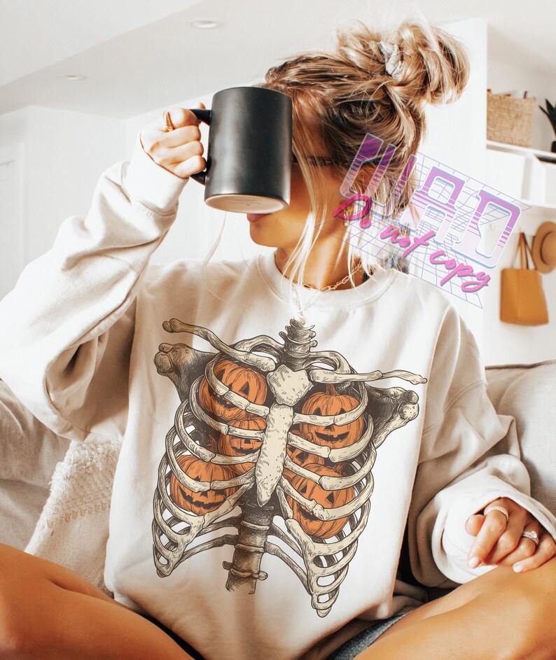May include: Cream-colored sweatshirt featuring a skeletal rib cage design with three carved orange pumpkins inside. The graphic is detailed with shading and highlights. The sweatshirt is a casual, long-sleeved garment.